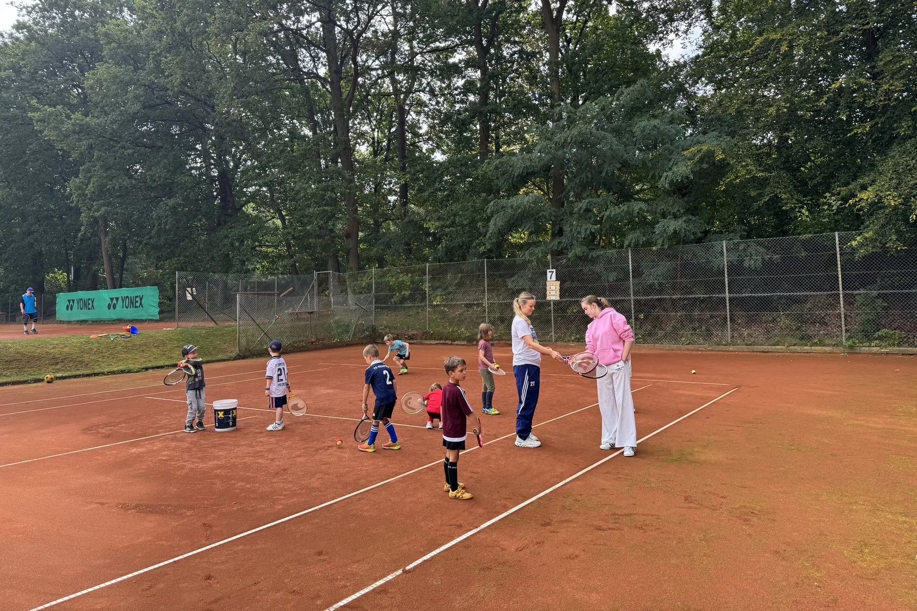 Kinder mit Tennisschlägern auf dem Sandplatz, zwei Trainerinnen zeigen den Griff